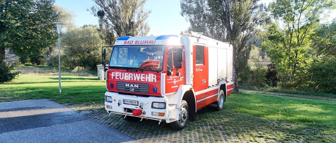 Ein rot-weißer Feuerwehrwagen mit 'Feuerwehr' und 'Bad Blumau' steht auf einem gepflasterten Bereich mit Rasen und Bäumen im Hintergrund.
