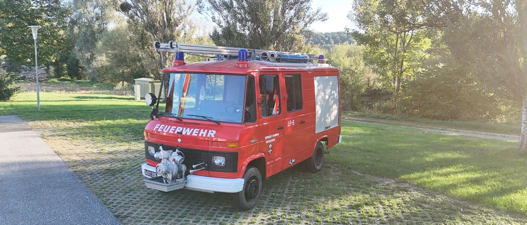Ein roter Feuerwehrwagen mit 'Feuerwehr' auf der Seite steht auf einer Kopfsteinpflasterstraße. Er hat eine Leiter auf dem Dach und eine Sirene auf dem Dach.
