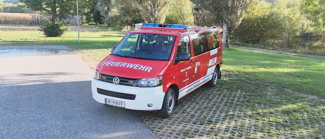 Ein rot-weißer VW-Bus mit 'Feuerwehr' auf der Seite ist auf einem gepflasterten Weg geparkt. Er hat ein blaues Licht oben und eine grüne Spielfigur auf der Windschutzscheibe.
