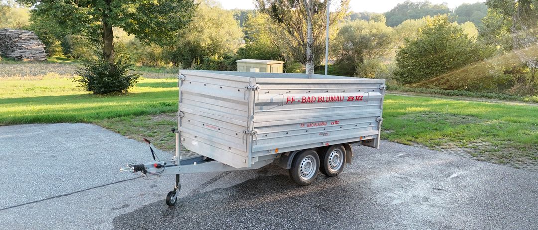 Ein silberner Anhänger mit der Aufschrift FF-BAD BLUMAI steht auf einem Kiesweg. Dahinter befindet sich eine Rasenfläche mit Bäumen und einem kleinen Gebäude.