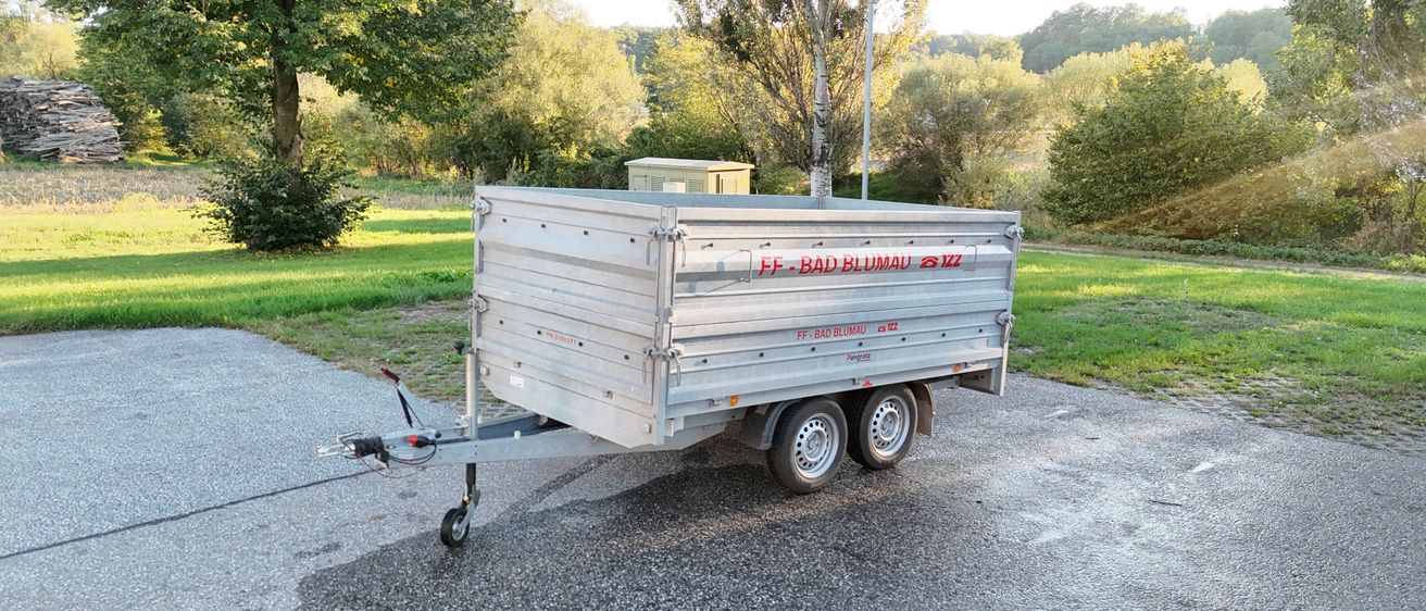 Ein silberner Anhänger mit der Aufschrift FF-BAD BLUMAI steht auf einem Kiesweg. Dahinter befindet sich eine Rasenfläche mit Bäumen und einem kleinen Gebäude.