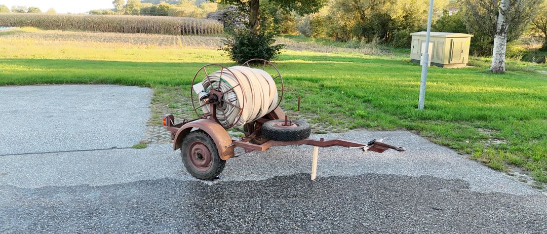 Ein alter Anhänger mit einem Reifen und einer Schlauchtrommel, der auf Kies neben einem Grasfeld geparkt ist.