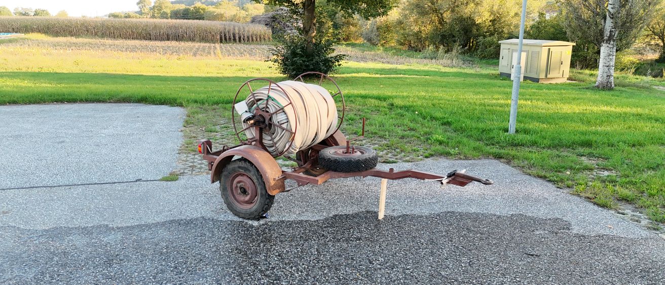 Ein alter Anhänger mit einem Reifen und einer Schlauchtrommel, der auf Kies neben einem Grasfeld geparkt ist.