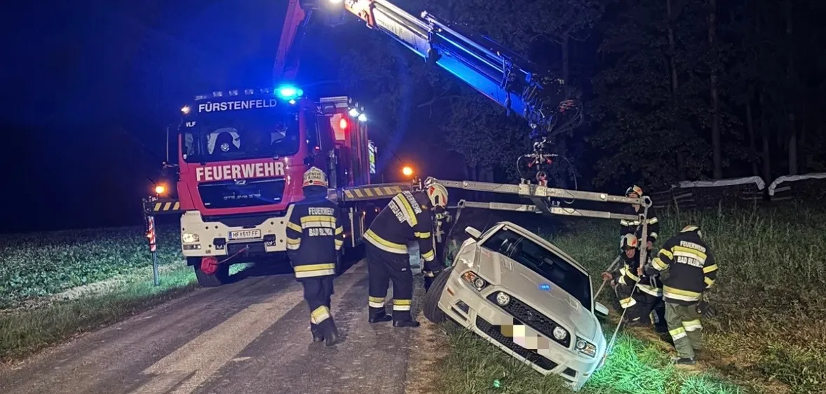 Ein Feuerwehrwagen steht nachts am Straßenrand. Ein Auto steht am Straßenrand und ein Kran hebt es. Einige Feuerwehrleute stehen um das Auto herum.
