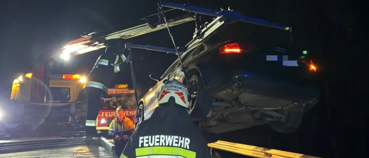 Ein Feuerwehrmann sichert ein Auto, das auf einen Flachbett-LKW gehoben wurde. Das Auto ist auf dem Kopf, und der Feuerwehrmann trägt einen Helm und eine reflektierende Weste.