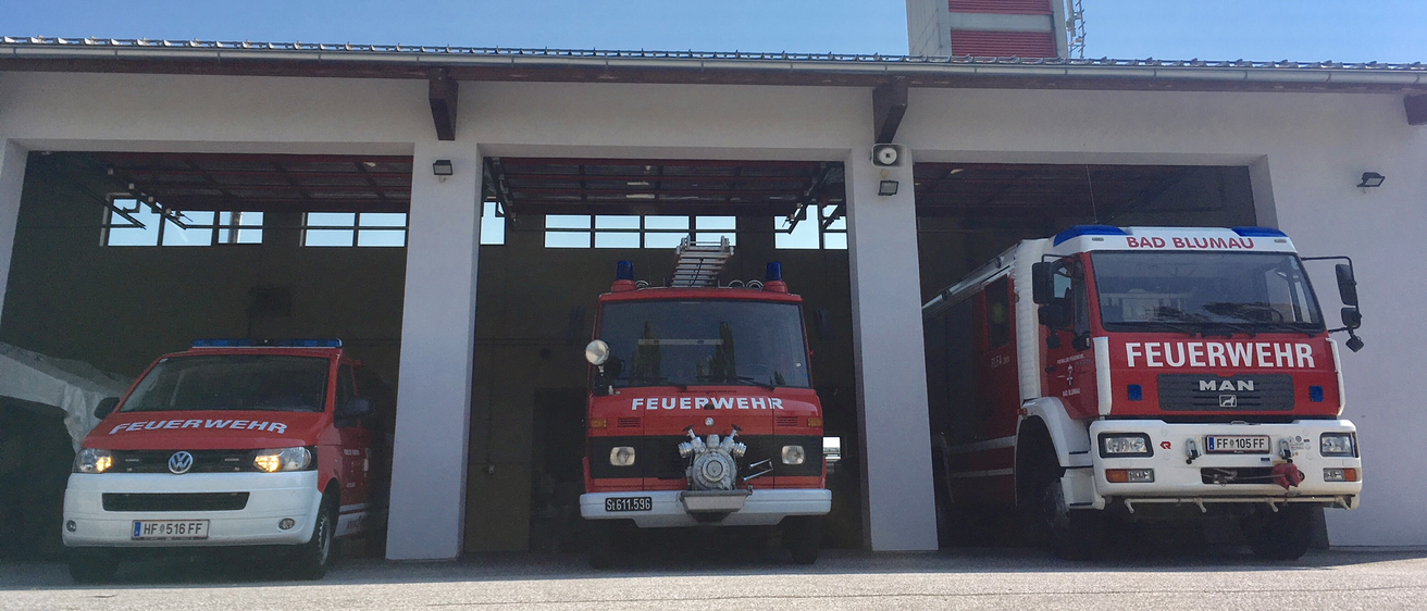 Drei rote Feuerwehrfahrzeuge sind in einer Garage unter blauem Himmel geparkt. Hinter der Garage steht ein Gebäude mit einem Turm.