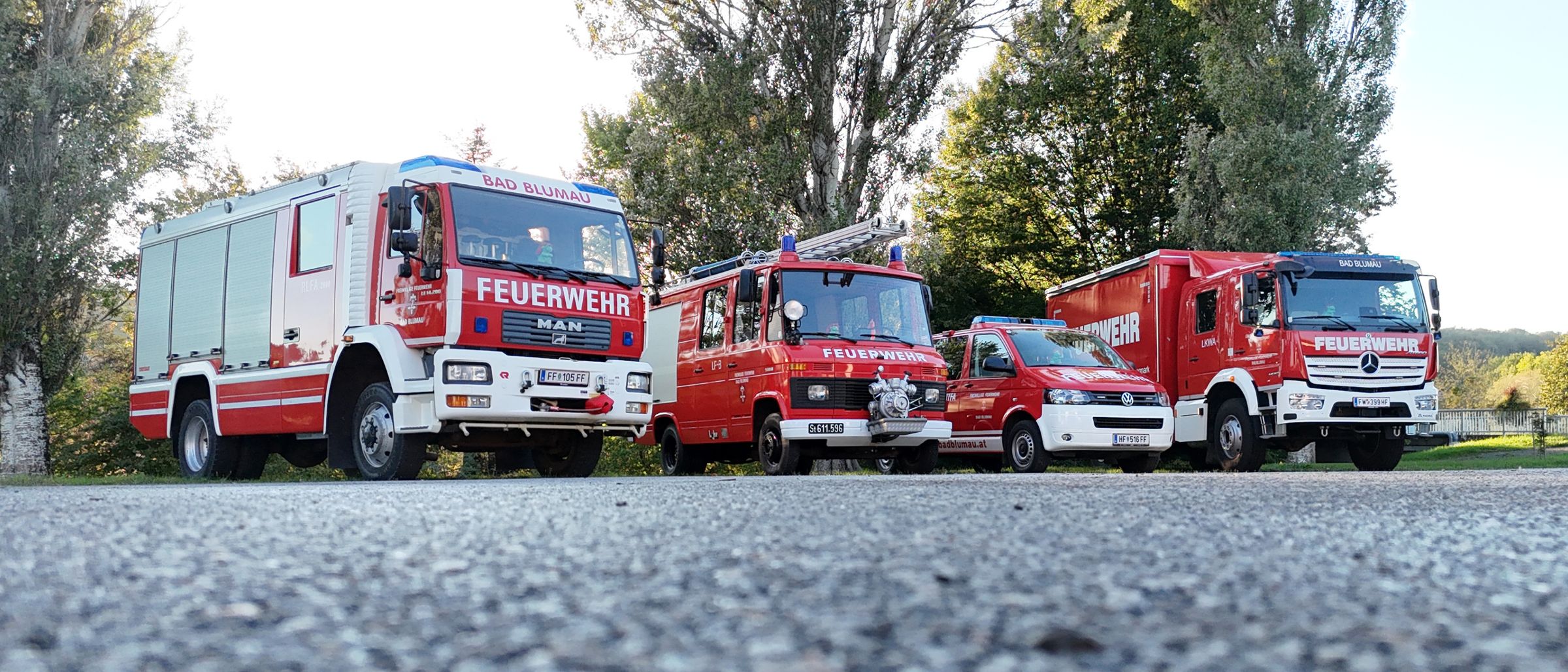 Vier rote Feuerwehrfahrzeuge sind auf einer Straße geparkt, mit Bäumen im Hintergrund. Jedes Fahrzeug hat das Wort 'Feuerwehr' an seiner Seite.