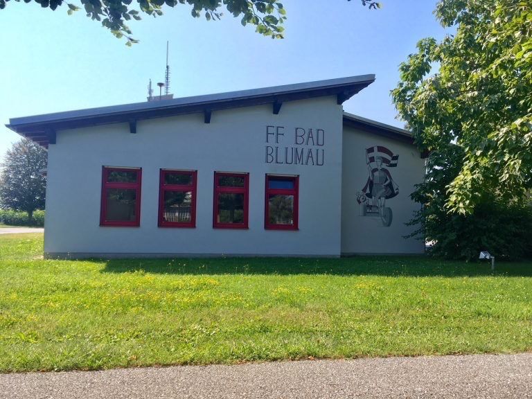 Ein weißes Gebäude mit roten Fenstern und einer Wandmalerei, die einen Ritter darstellt, mit der Aufschrift FF BAD BLUMAu, in einem grasbewachsenen Bereich unter einem klaren Himmel.
