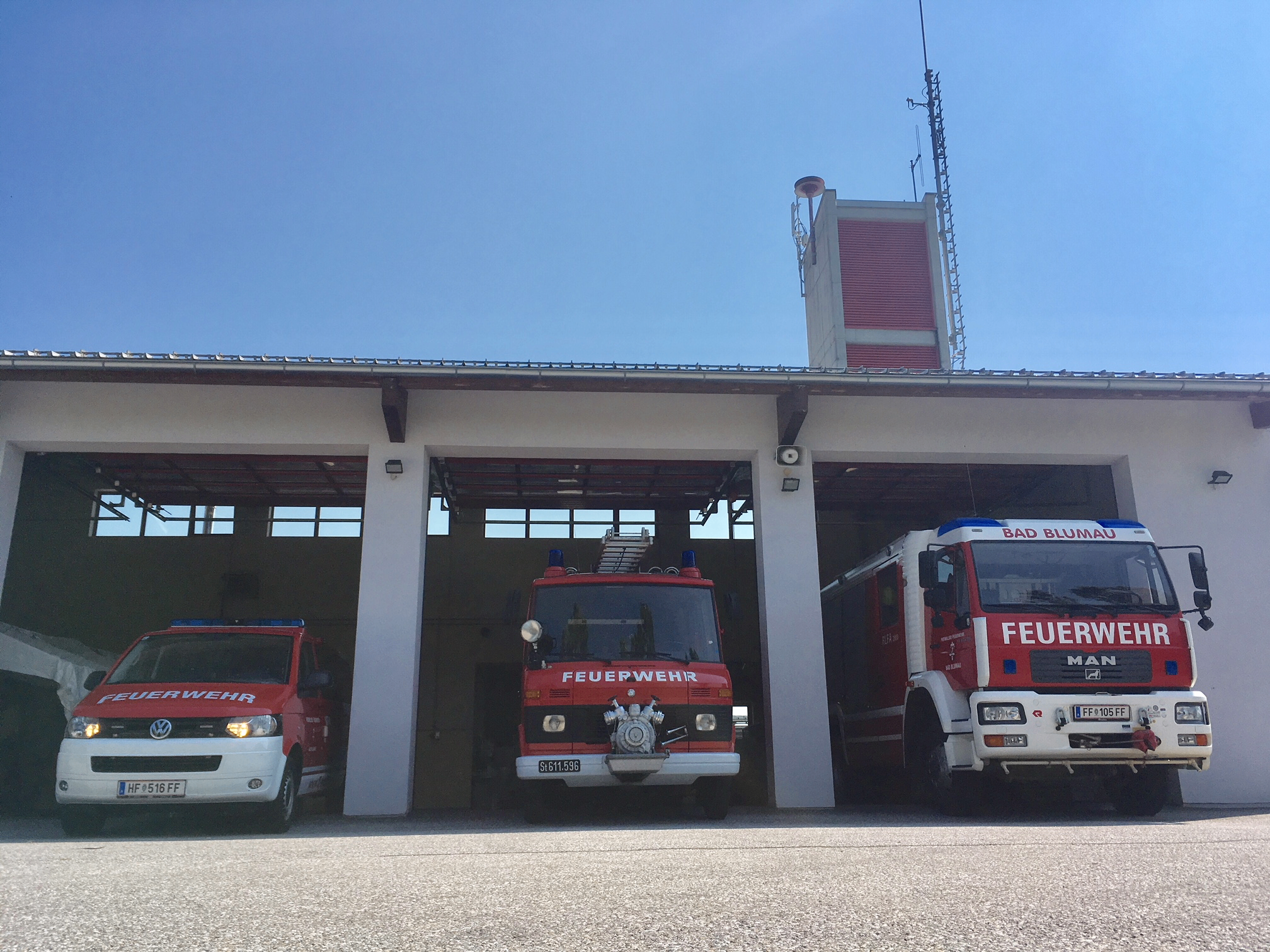 Drei Feuerwehrfahrzeuge sind in einer Feuerwache unter einem klaren blauen Himmel geparkt. Das mittlere Fahrzeug hat 'Feuerwehr' darauf geschrieben.