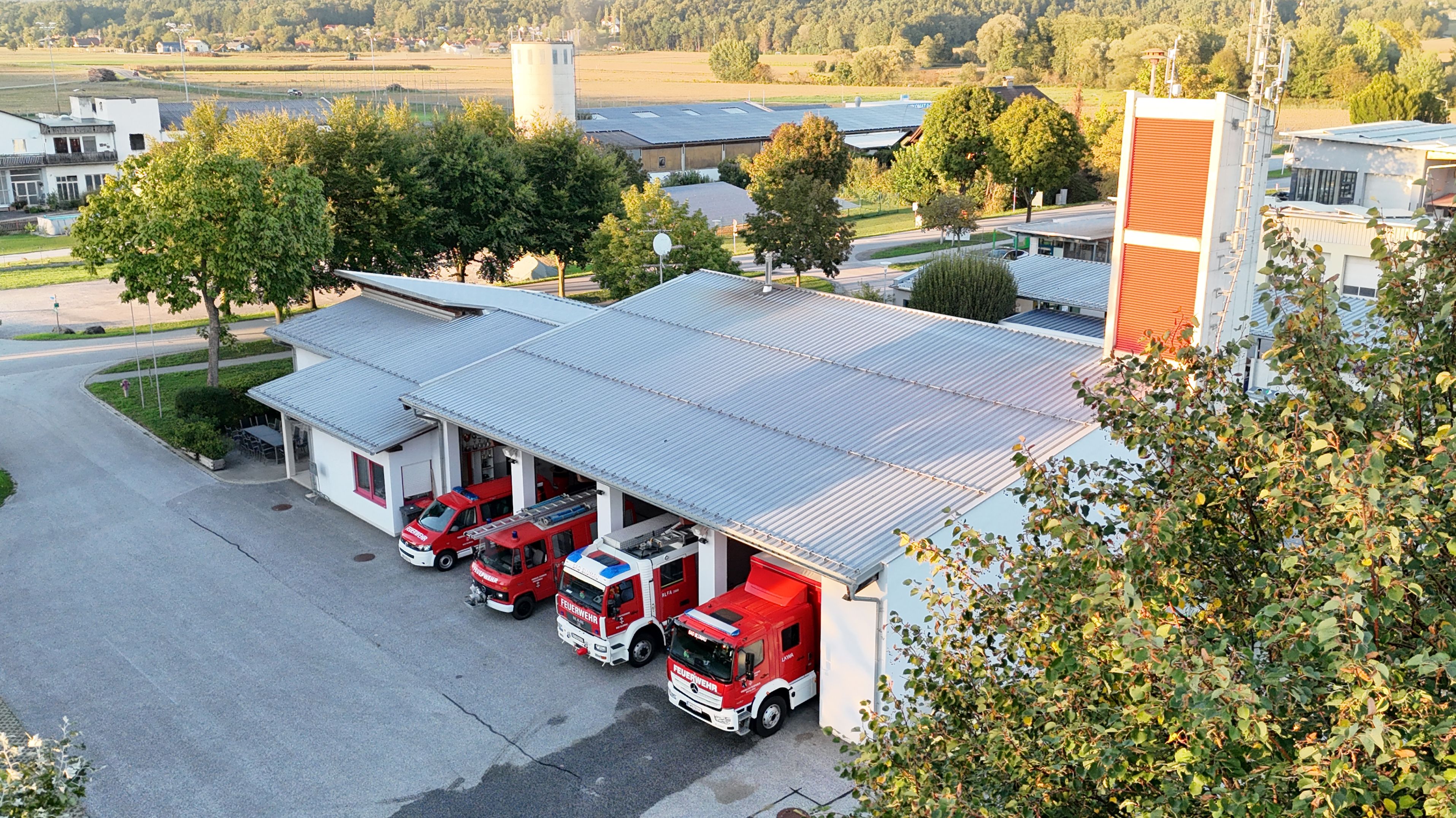 Ein Blick von oben auf eine Feuerwehrstation mit drei roten Feuerwehrfahrzeugen vor dem Gebäude. Im Hintergrund sind Bäume und ein Silo zu sehen.