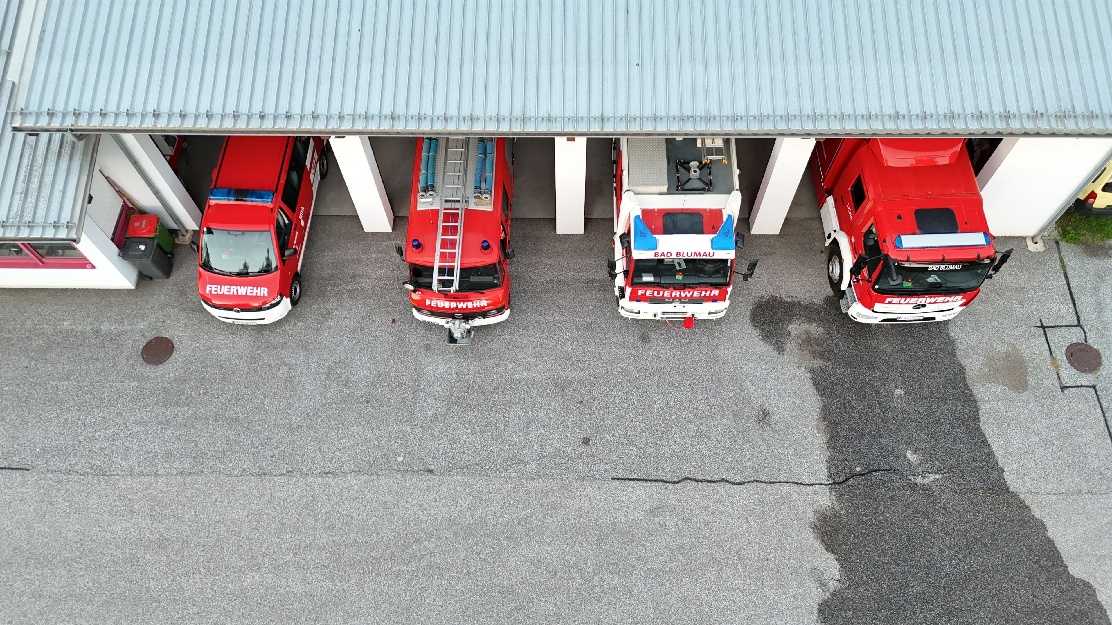 Eine Draufsicht auf eine Feuerwache mit vier roten Feuerwehrfahrzeugen, die in einer Reihe unter einem grauen Dach geparkt sind.