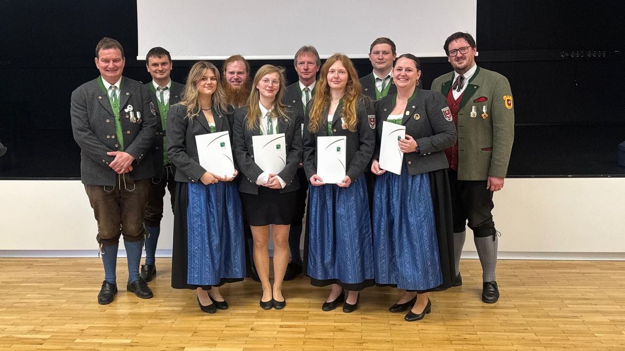 Eine Gruppe von Menschen in traditioneller deutscher Kleidung steht zusammen, hält Urkunden in der Hand und lächelt für ein Foto auf einem Holzboden.