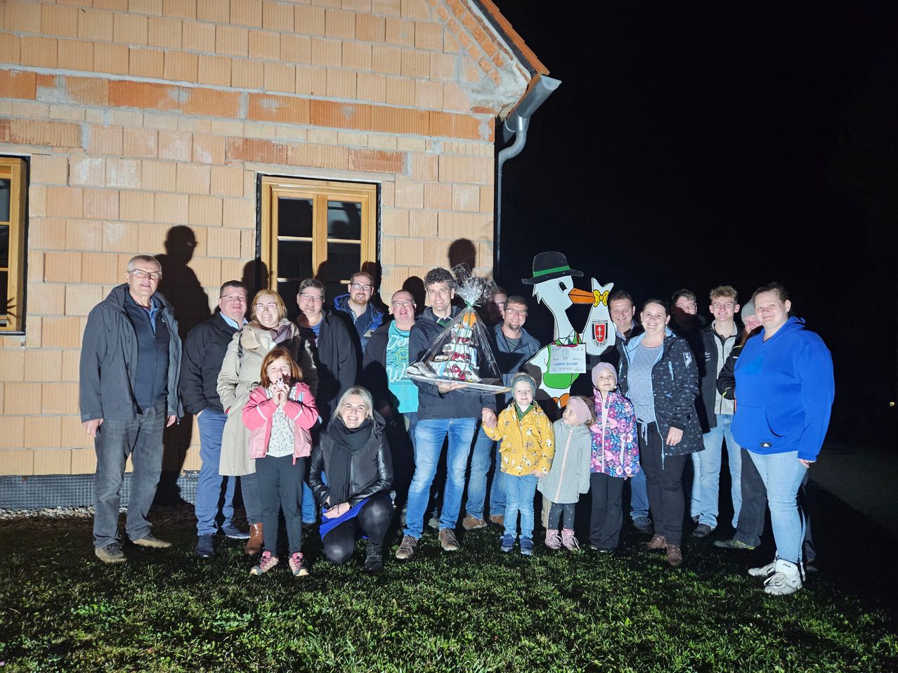 Eine Gruppe von Erwachsenen und Kindern posiert für ein Foto vor einem Haus bei Nacht, ein Mann hält eine Geschenktüte, ein Storch-Ausschnitt steht im Hintergrund.