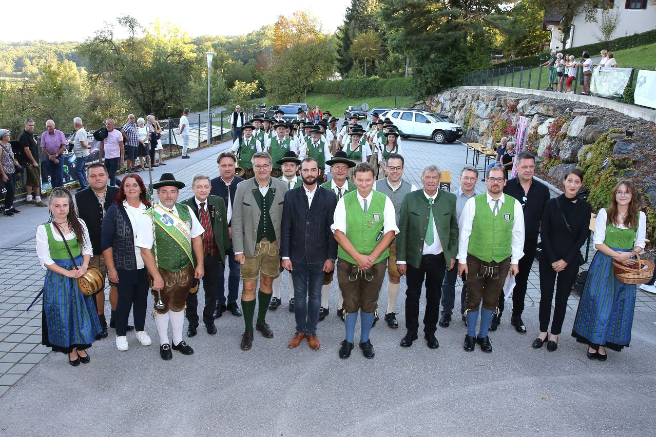 Eine Gruppe von Menschen in traditioneller deutscher Kleidung, die vor einer Steinmauer mit Bäumen und geparkten Autos steht. Sie posieren für ein Foto mit einem Mann im Anzug in der Mitte.