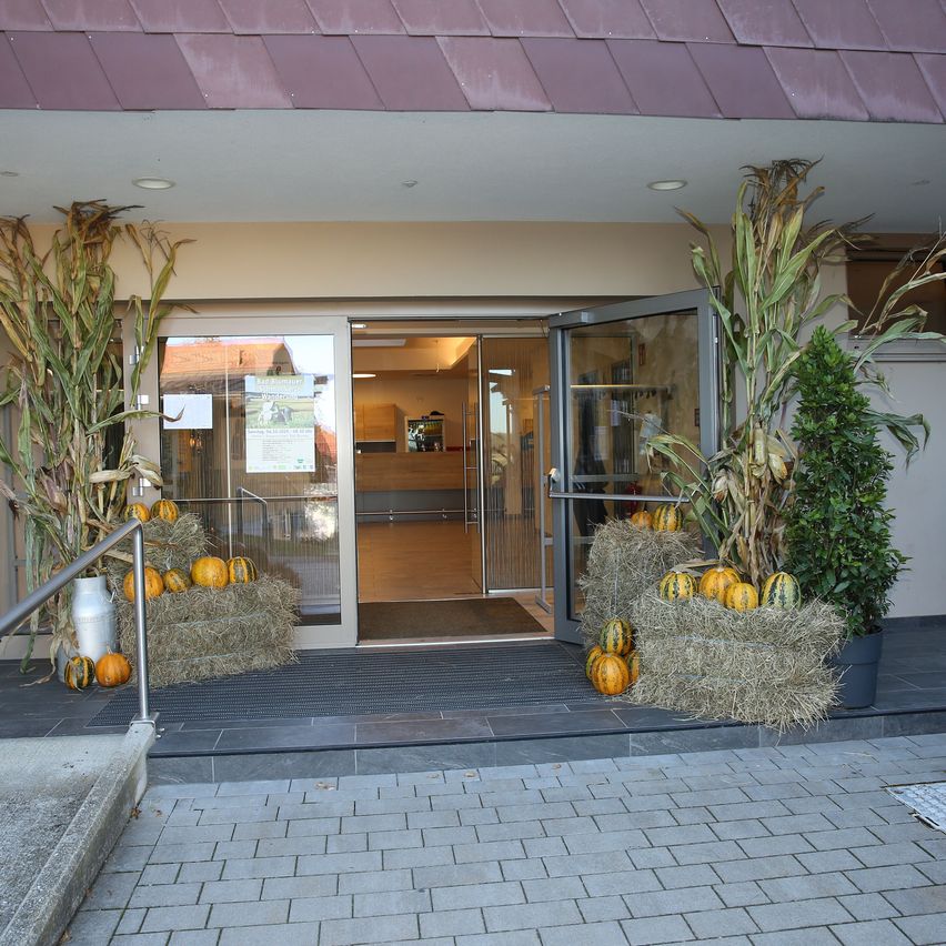 Der Eingang eines Gebäudes ist mit Heuballen, Kürbissen und getrocknetem Mais dekoriert. Die Türen sind offen und zeigen einen Flur. Ein Plakat ist an der Glastür angebracht.
