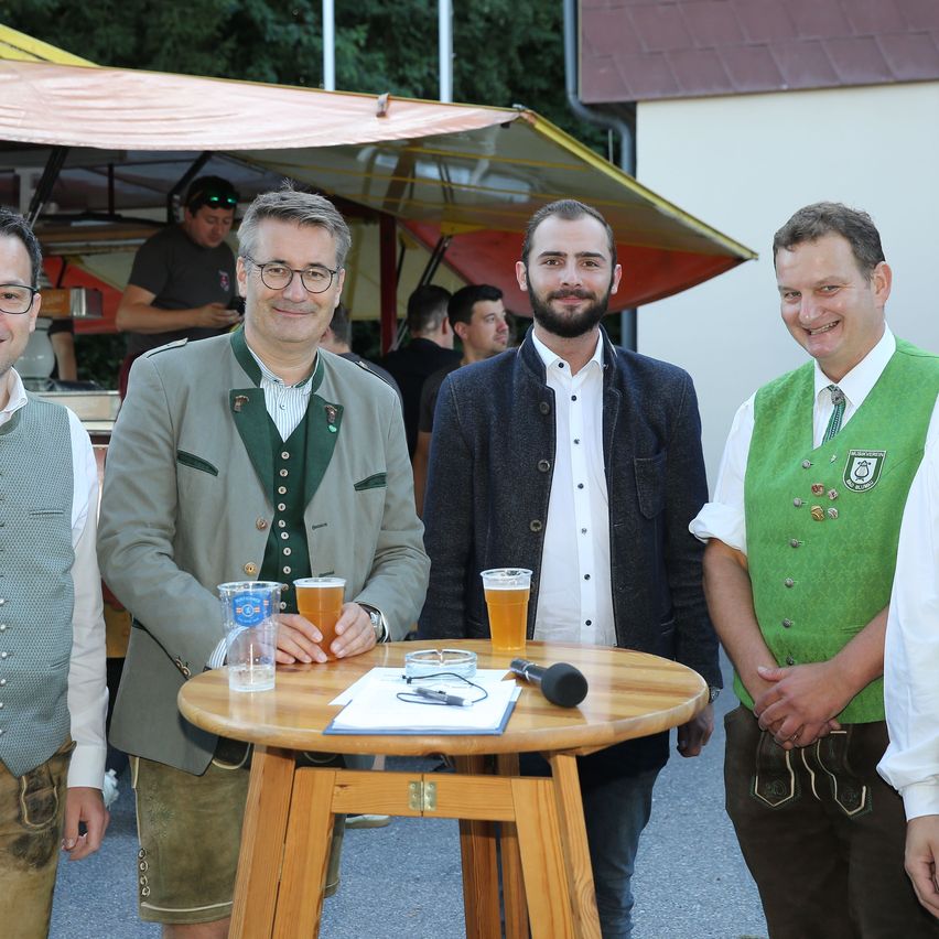 Vier Männer in traditioneller Kleidung posieren für ein Foto. Sie stehen um einen kleinen Holztisch mit Biergläsern und einem Mikrofon. Die Umgebung scheint ein Outdoor-Event zu sein.