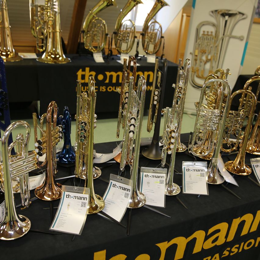 Ein in Schwarz gedeckter Tisch zeigt verschiedene Blechblasinstrumente, darunter Trompeten, Posaunen und Tuben. Die Instrumente sind in verschiedenen Farben und einige haben weiße Etiketten. Der Hintergrund ist gelb und schwarz.