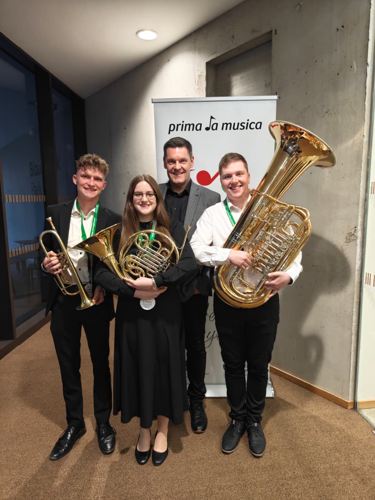 Vier Personen posieren mit ihren Musikinstrumenten für ein Foto. Hinter ihnen ist ein Banner mit den Worten 'prima la musica.'