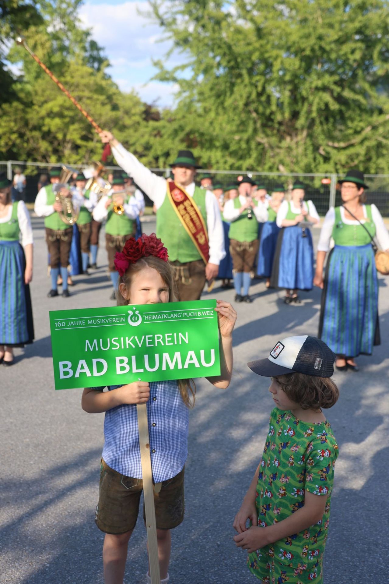 Ein junges Mädchen hält ein grünes Schild mit der Aufschrift 'Musikverein Bad Blumau', während sie vor einer Blaskapelle in traditioneller deutscher Kleidung steht.