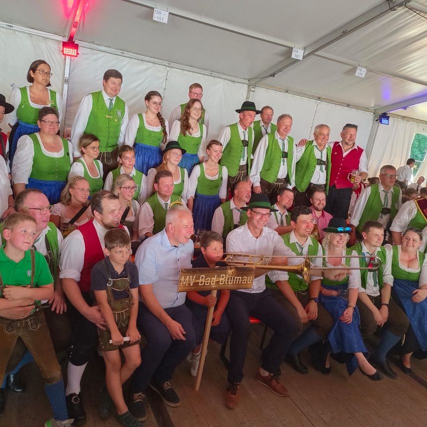 Eine Gruppe von Menschen in traditioneller deutscher Kleidung, einschließlich grüner Westen und Hüten, versammelt sich in einem Zelt für ein Foto. Einige halten Musikinstrumente, und ein Schild steht 'MV Bad Blumau.'