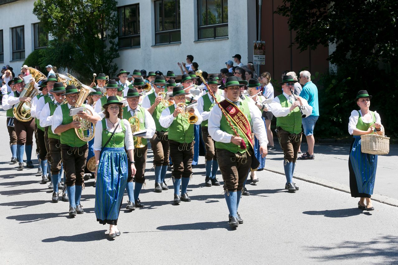 Eine Gruppe von Menschen in traditioneller bayerischer Kleidung marschiert bei einem Umzug und spielt verschiedene Musikinstrumente.