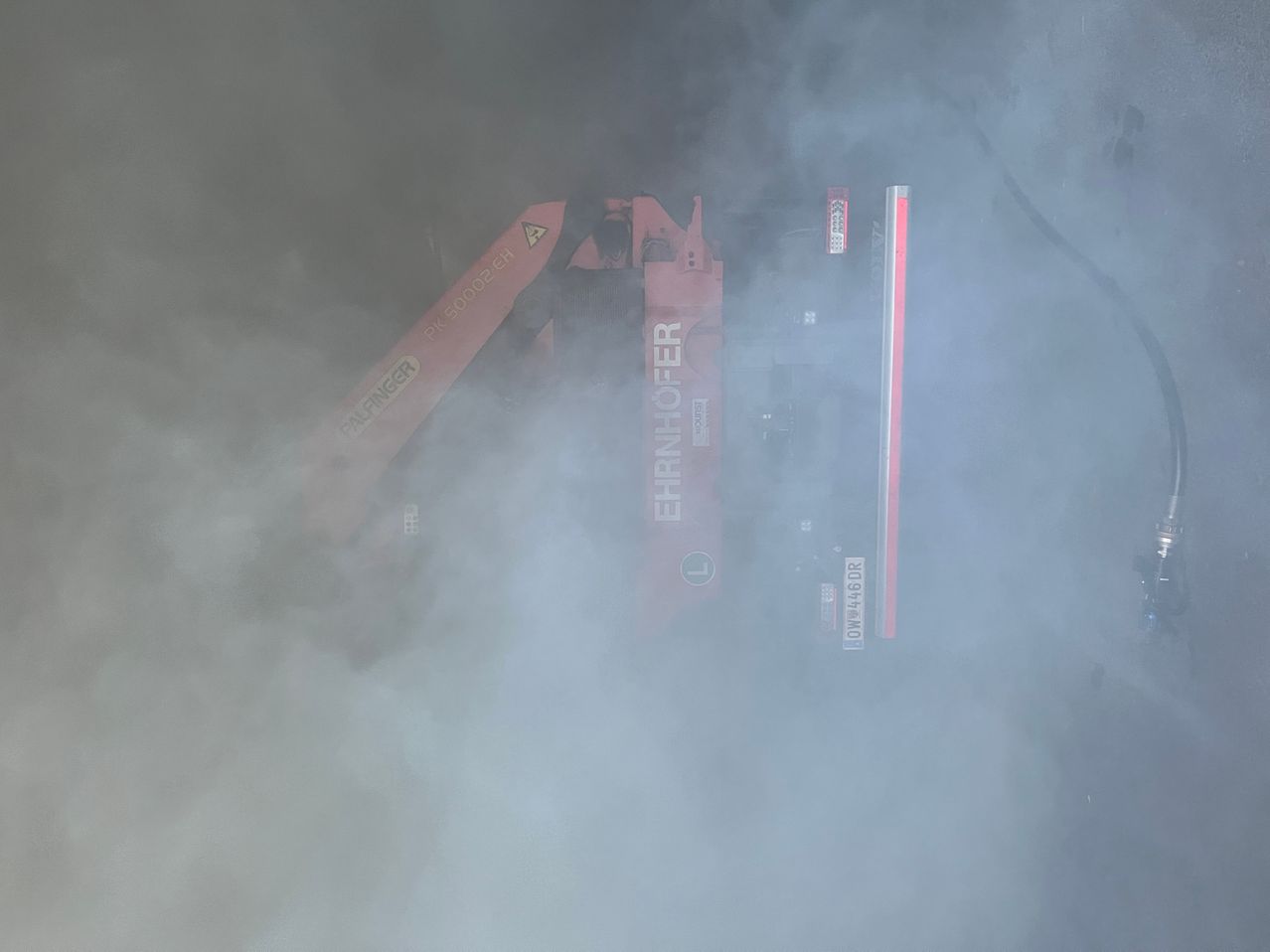 Ein roter Baufahrzeug ist teilweise durch Rauch verdeckt, wobei Teile mit 'Ehrlichhofer' und 'L' sichtbar sind. Der Rauch ist dick und weiß.