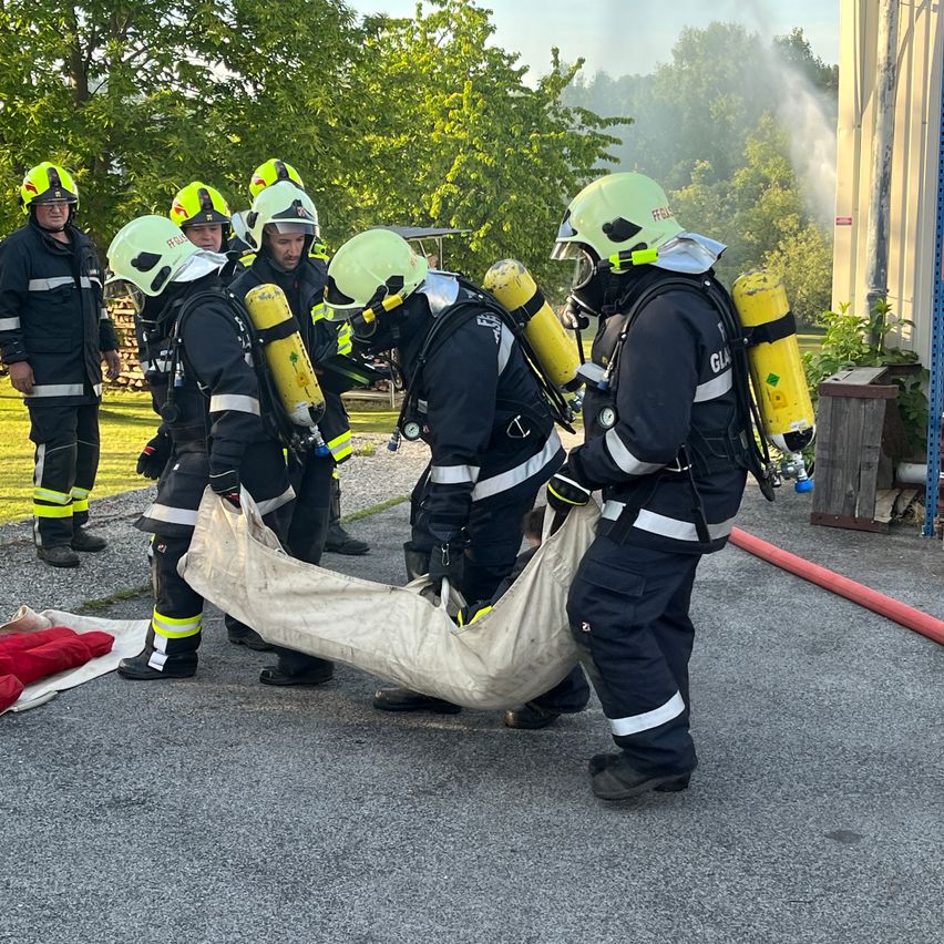 Feuerwehrleute in voller Ausrüstung üben Rettungsaktionen mit einer Puppe auf einer asphaltierten Oberfläche. Ein Team hält eine große Tasche, während ein anderes einen Feuerwehrschlauch hält. Bäume und ein Gebäude sind im Hintergrund.