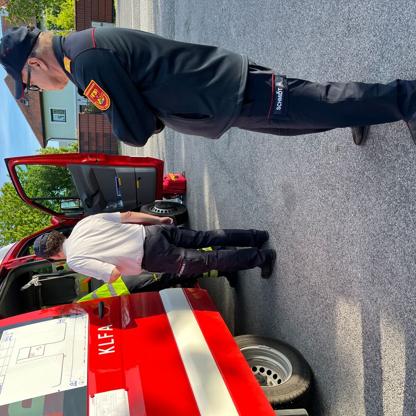 Zwei Feuerwehrleute arbeiten an einem roten Fahrzeug. Einer liegt auf dem Boden und überprüft etwas, während der andere steht und beobachtet.