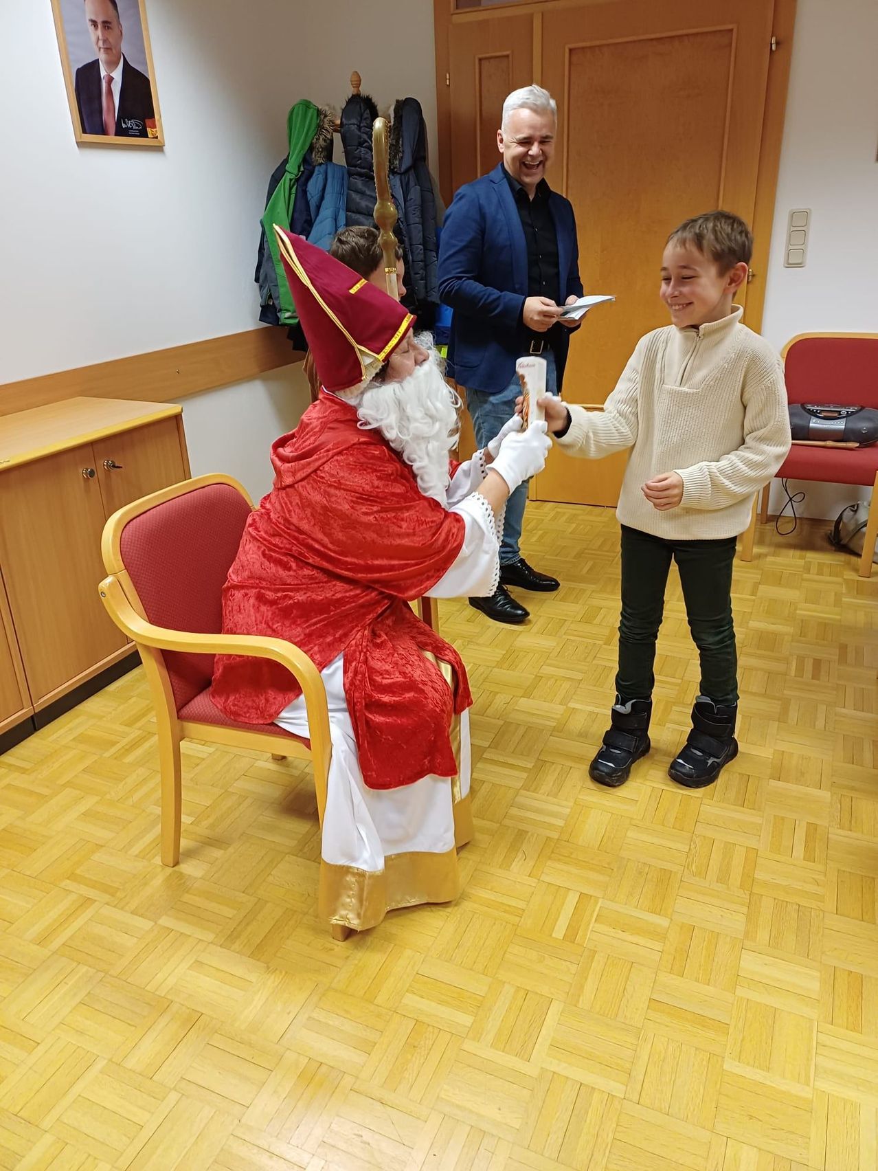 Ein Junge erhält ein Geschenk von einer Person, die als Weihnachtsmann verkleidet ist. Ein anderer Mann steht in der Nähe und hält ein Papier. Im Hintergrund befinden sich ein Holzschrank und ein Stuhl.