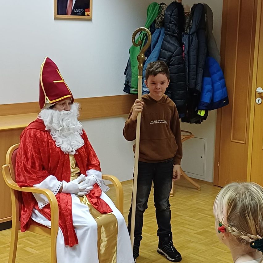 Eine Person in der Kleidung des Heiligen Nikolaus sitzt auf einem Stuhl, während ein Junge einen Stab hält. Sie befinden sich in einem Raum mit Holzboden und Wintermänteln an der Wand.