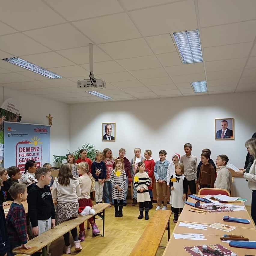 Kinder versammeln sich in einem Klassenzimmer, vor einem langen Tisch stehend. Sie sind in Winterkleidung gekleidet und einige tragen gelbe Sterne. Eine Frau steht rechts und hält ein Papier. Hinter ihnen hängen zwei gerahmte Porträts an der Wand.