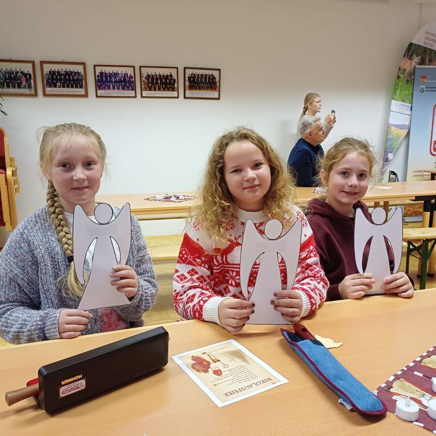 Drei Mädchen posieren mit engelsförmigen Ausschnitten, sitzend an einem Tisch mit verschiedenen Gegenständen, darunter ein Holzwalze, ein Beutel und ein Stück Papier. Dahinter steht eine Person und macht ein Foto. Der Hintergrund zeigt gerahmte Bilder an der Wand.