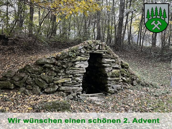 Eine Steinhütte steht in einer Waldlichtung, umgeben von gefallenen Blättern. Der Eingang ist dunkel. Ein grünes Emblem mit Bäumen und einem Hammer ist rechts. 'Wir wünschen einen schönen 2. Advent' steht unten geschrieben.