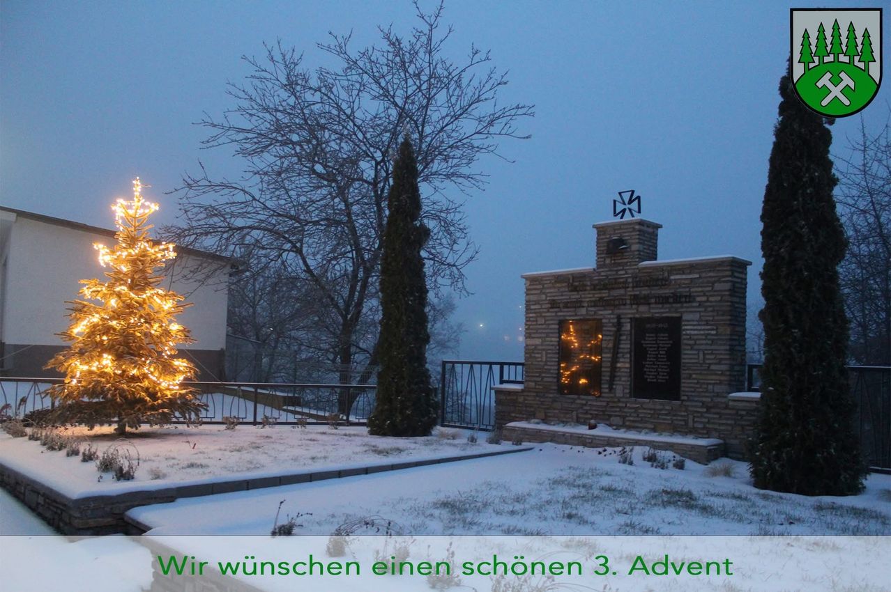 Eine verschneite Szene mit einem beleuchteten Weihnachtsbaum, einem kahlen Baum und einem Gedenkstein mit einem Kreuz. Es befindet sich auf einem Hügel mit Geländern. Im Hintergrund ist ein Gebäude sichtbar.