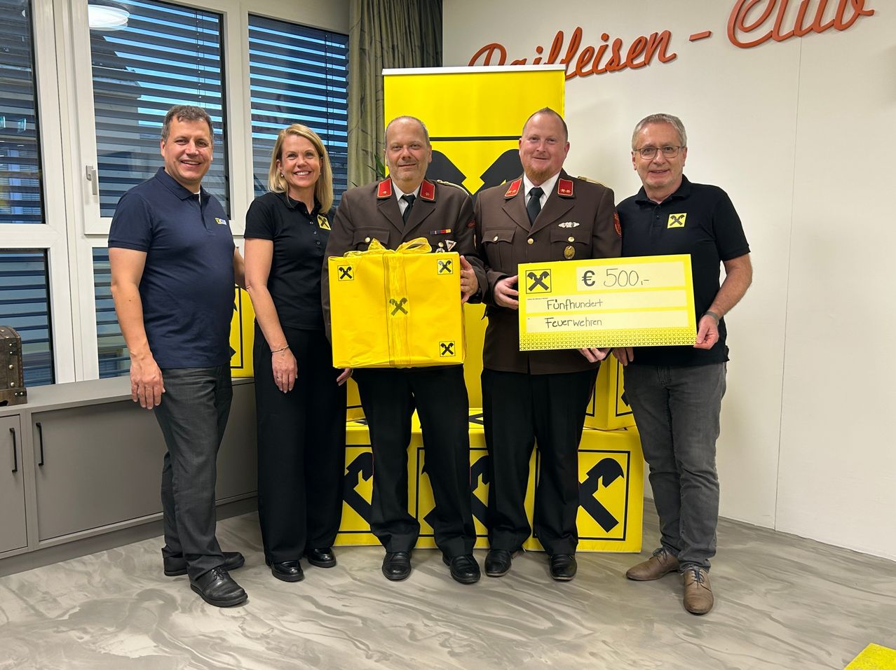 Fünf Personen stehen in einem Raum mit gelben Kisten und einem Banner. Zwei Männer in Uniform halten einen Scheck und eine gelbe Tasche. Die anderen lächeln.