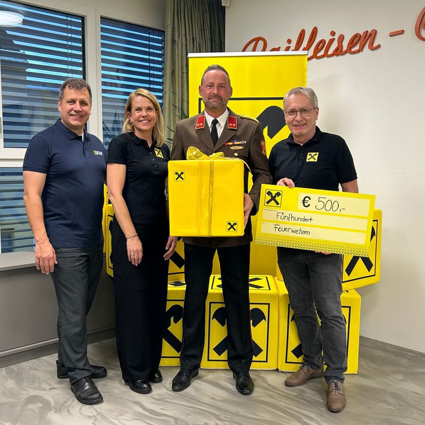 Vier Personen stehen vor gelben Boxen und lächeln. Ein Mann in Uniform hält eine Geschenkbox und ein Zertifikat mit einem Geldbetrag. Der Ort ist wahrscheinlich ein Büro oder Studio.