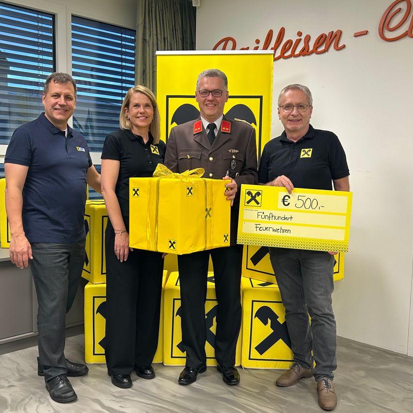 Vier Personen posieren vor gelben Boxen, zwei Männer in Schwarz und eine Frau in Gelb, die eine gelbe Box und einen Scheck über 500 Euro hält. Dahinter ist ein gelber Banner mit Text und Symbolen angebracht.