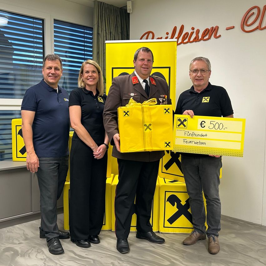 Vier Personen stehen vor gelben Boxen; die zentrale Figur hält eine gelbe Tasche und einen Scheck über 500 Euro. Dahinter steht ein Schild mit der Aufschrift 'Dilleisen-C'.