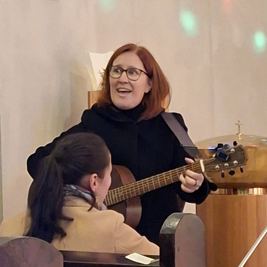 Eine Frau mit Brille spielt lächelnd eine Gitarre, während ein junges Mädchen vor ihr in einem Raum mit weißen Wänden zuhört.