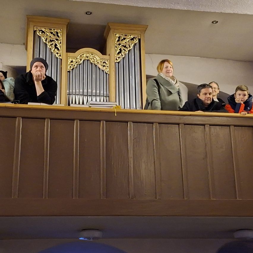 Eine Gruppe von Menschen steht auf einer Galerie und beobachtet eine Veranstaltung. Ein Mann trägt eine Mütze und bedeckt seinen Mund. Hinter ihnen steht eine große Orgel.