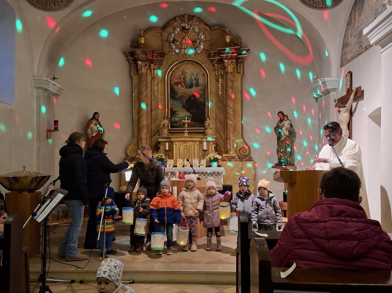 Mehrere Kinder halten Laternen und stehen vor einem Altar einer Kirche mit einem Gemälde. Hinter ihnen steht eine Gruppe von Erwachsenen. Darüber leuchtet ein Diskolicht.