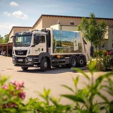 Ein weißer Lkw mit einer grünen Werbung für 'So Action' steht vor einem Gebäude mit mehreren Fenstern und einem roten Auto, das in der Nähe geparkt ist.
