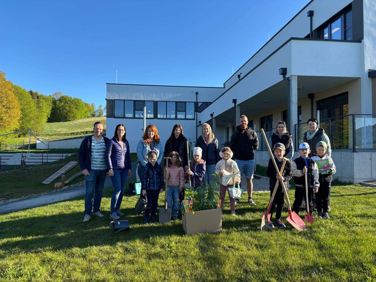 Eine Gruppe von Erwachsenen und Kindern posiert für ein Foto vor einem Gebäude mit einem Topf. Sie halten Schaufeln und Werkzeuge.