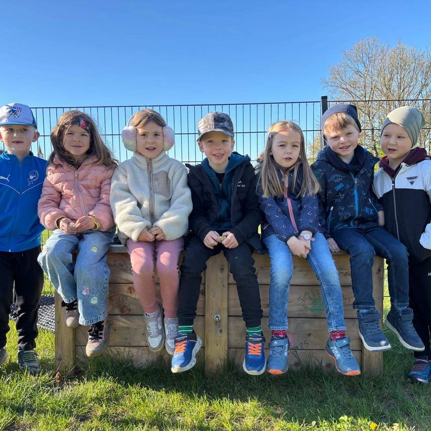 Eine Gruppe von Kindern sitzt auf einer Holzbank im Park. Sie lächeln und scheinen für ein Foto zu posieren. Dahinter befindet sich ein schwarzer Zaun und ein Baum.