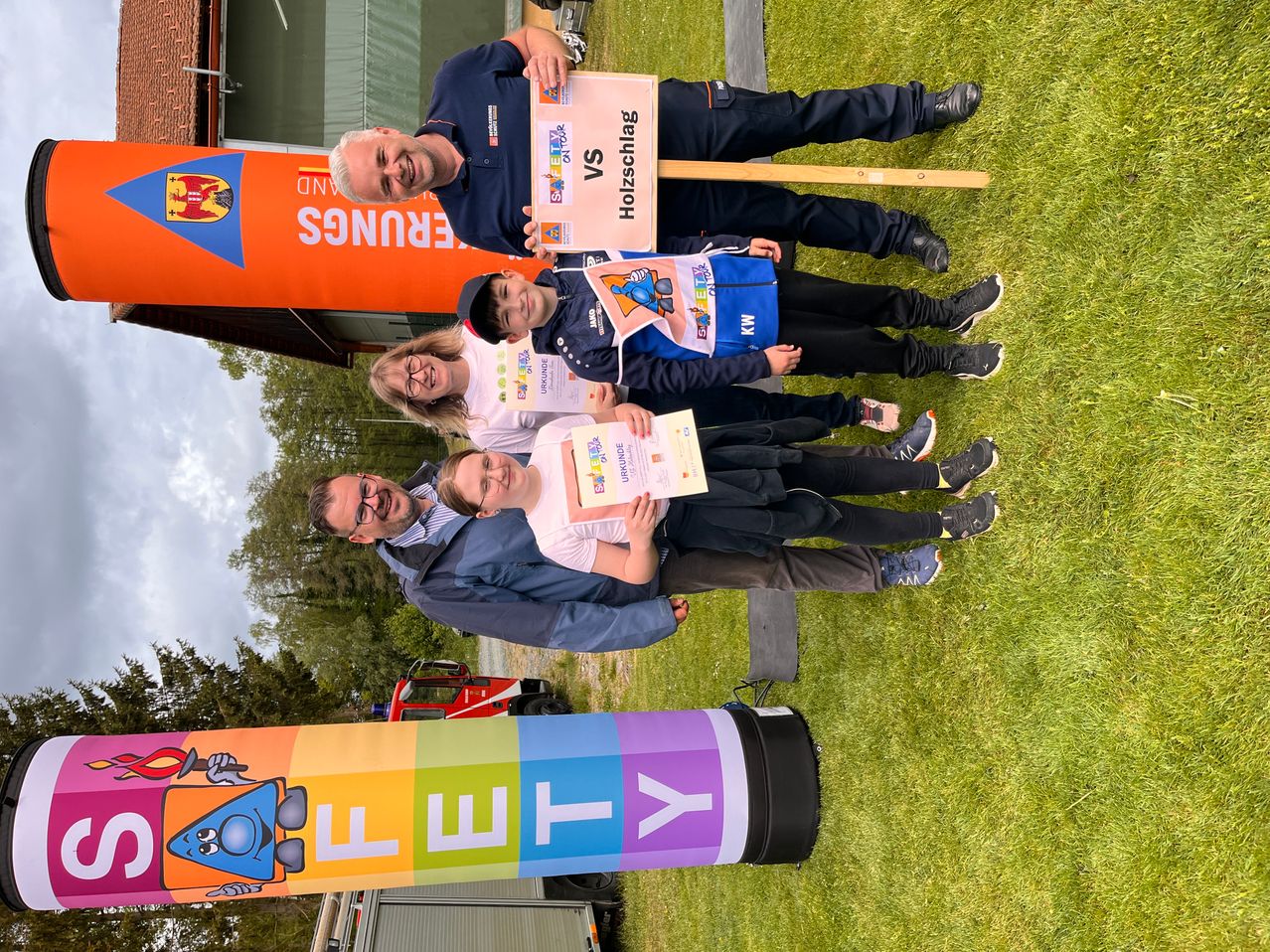 Eine Gruppe von Erwachsenen und Kindern steht lächelnd für ein Foto auf dem Rasen. Ein Mann hält ein Schild mit der Aufschrift Holzschlag VS. Die Kinder halten Urkunden in der Hand. Bunte Banner mit Buchstaben stehen hinter ihnen.