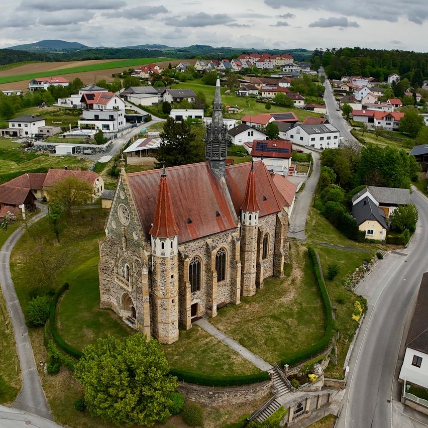 Luftaufnahme einer mittelalterlichen Kirche mit roten Dächern, umgeben von Häusern, Straßen und üppigem Grün. Die Kirche hat einen Turm und Türme mit einer klaren blauen Himmel darüber.