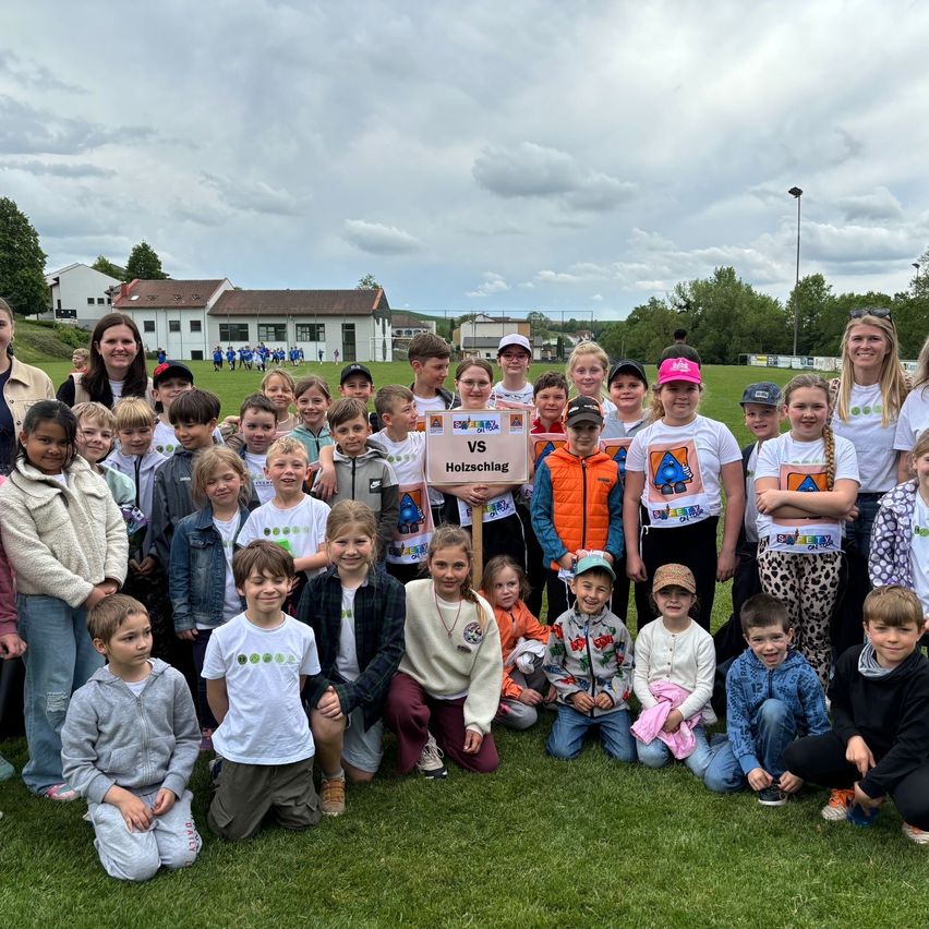 Eine Gruppe von Kindern und Erwachsenen posiert auf einem grünen Feld für ein Foto mit einem Banner, das 'VS Hotzschlag' steht. Einige tragen weiße T-Shirts mit grünen Symbolen. Bäume und ein Gebäude sind im Hintergrund.