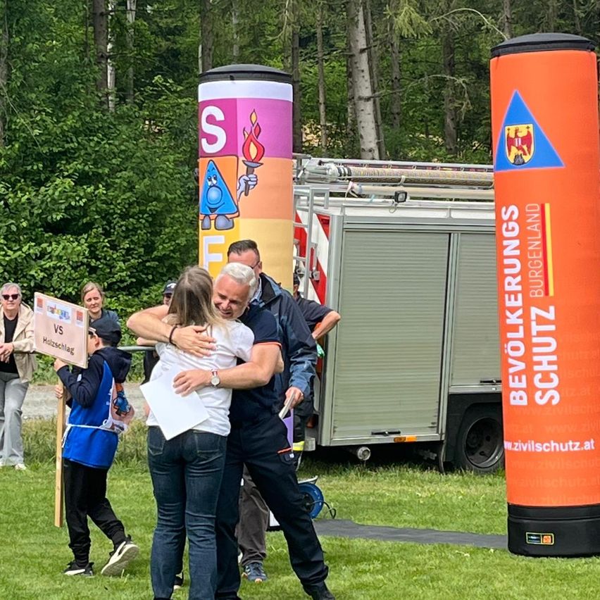 Eine Gruppe von Menschen versammelt sich auf einem Rasenfeld. Zwei Personen umarmen sich, während andere im Hintergrund Schilder halten. Ein Feuerwehrwagen ist im Hintergrund geparkt.