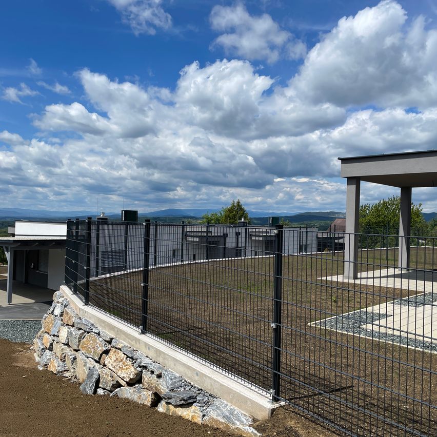 Eine Baustelle mit Steinmauer und schwarzem Zaun. Der Himmel ist klar mit einigen Wolken.