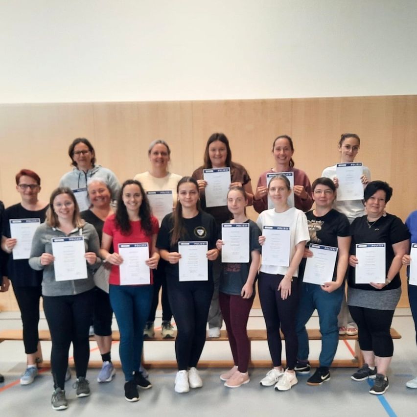 Eine Gruppe von Frauen steht in einem Fitnessstudio und hält Urkunden in der Hand. Sie tragen Freizeitkleidung, und einige haben eine Brille. Der Hintergrund ist eine Holzwand. Sie lächeln für ein Foto.